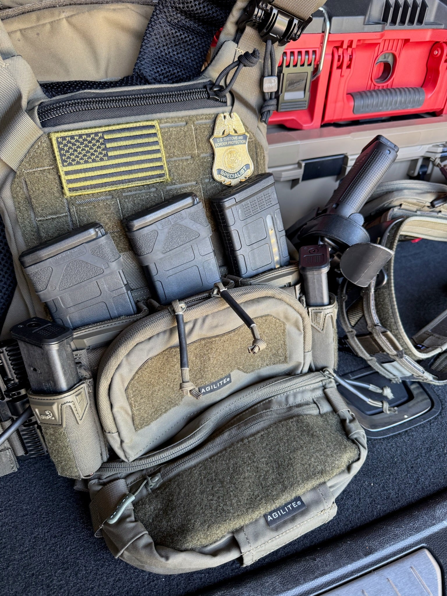 Close-up of tactical flex badge for a CBP Agent and gear with pouches and American flag patch on a vehicle interior.